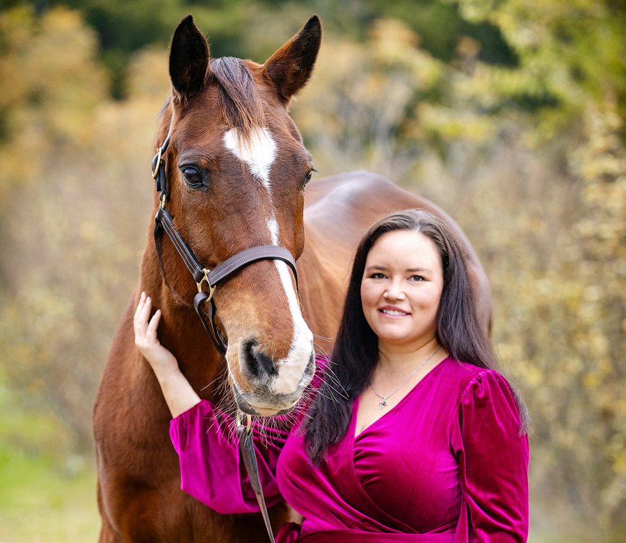 Jessica smiling beside her chestnut horse, who wears a leather halter, with autumn trees behind them
